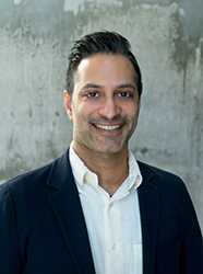 A smiling man with dark hair wearing a dark blazer over a white button-down shirt against a gray wall.