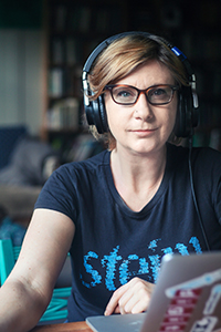 A woman with short blonde hair, glasses, and large headphones sits at a desk with a laptop.