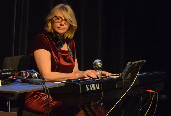 A woman with blonde hair and glasses playing a black Kawai electronic keyboard in a dark room.