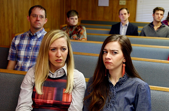 Four people with somber expressions sit in rows of wooden pews.