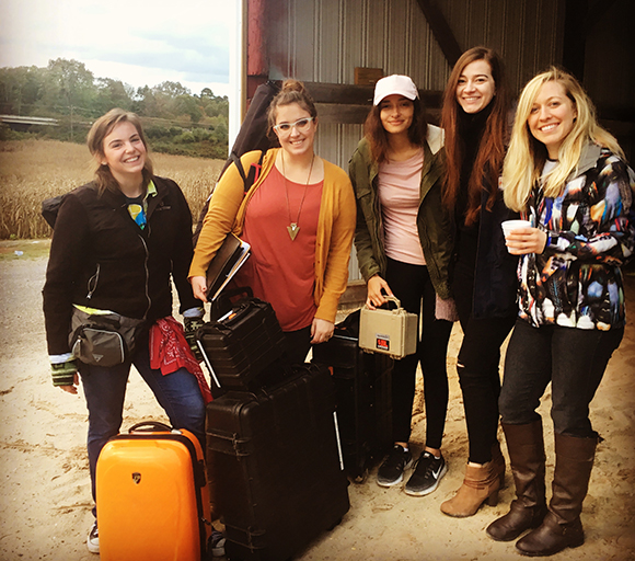 Five smiling women stand outdoors with suitcases, a hard case, and a backpack near a barn.