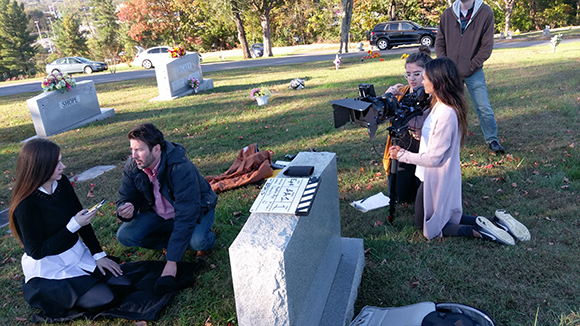 A film crew with a camera and clapperboard filming two actors sitting on the grass in a cemetery.