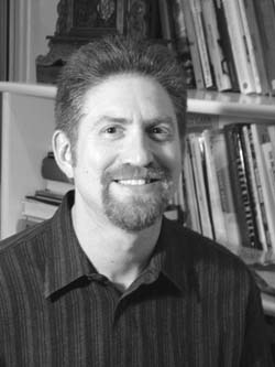 A smiling man with a goatee wearing a striped shirt, positioned in front of bookshelves.