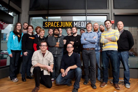 A diverse group of people posing for a group photo in an office setting in front of a Spacejunk Media sign.