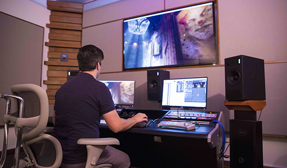 A man sits at a desk with multiple monitors, studio speakers, and a mixing console in a darkened studio.