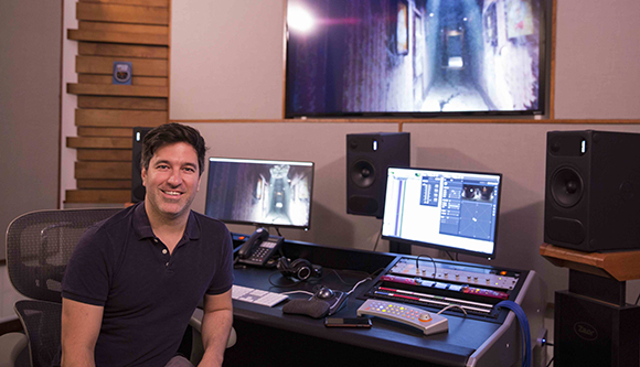 A man smiling while sitting at a desk with computer monitors, studio speakers, and audio mixing equipment.
