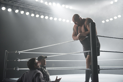 A muscular man leans over wrestling ring ropes looking down at two men in a dimly lit arena.