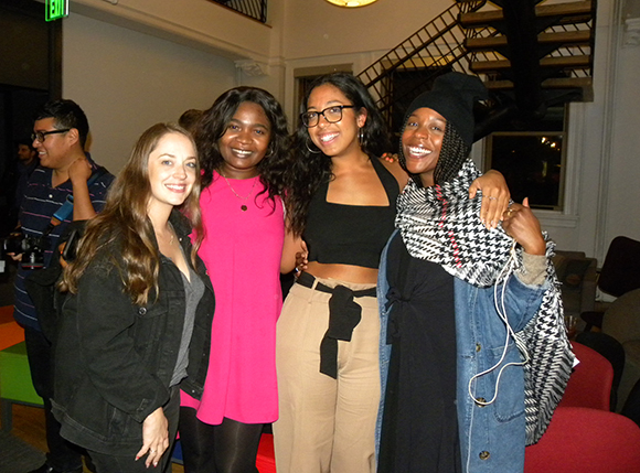 Four smiling women posing together indoors near a staircase.