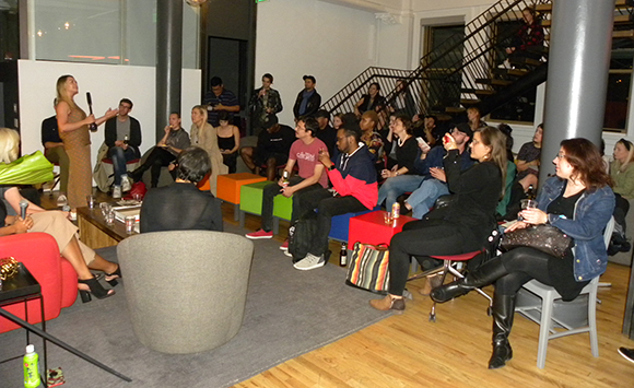 A woman in a gold dress stands speaking to a diverse group of people seated on chairs and colorful ottomans.