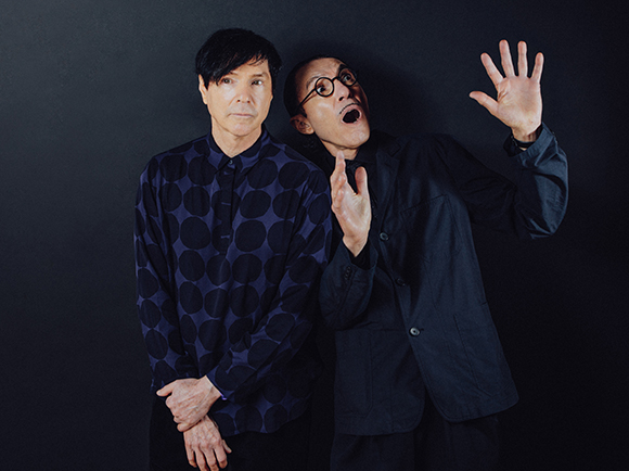 Two men in dark clothing pose against a black background; one looks serious while the other has an expressive face.