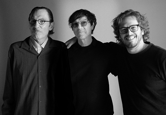Three men wearing glasses and dark clothing pose together in a black and white portrait.