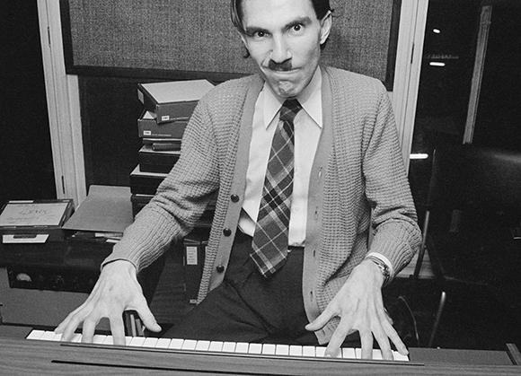 Man in a cardigan and plaid tie playing a keyboard with a wide-eyed, intense facial expression.