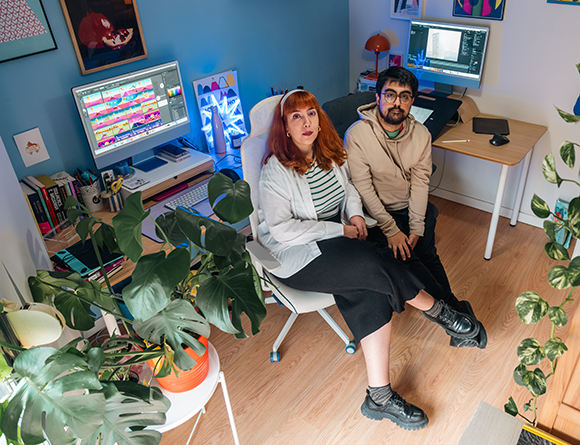 A man and woman sit together in a bright home office surrounded by indoor plants and dual computer setups.