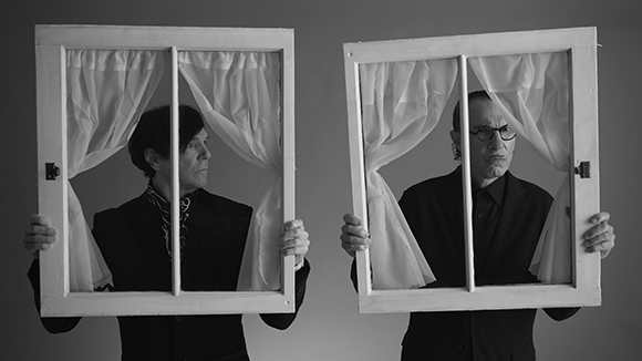 Two men in dark suits hold white window frames with curtains in front of their faces against a gray background.
