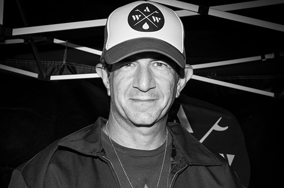 A man in a trucker hat and jacket smiling in front of a dark background.