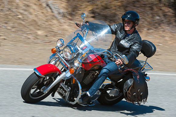 A man in a black leather jacket and helmet rides a red cruiser motorcycle around a curve.