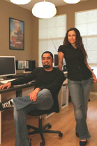 A man sits in an office chair while a woman stands beside him in a room with computers.