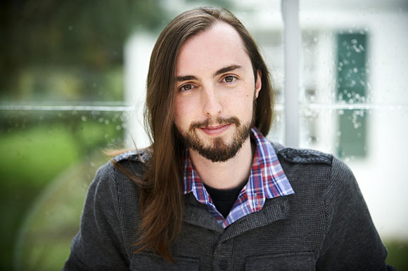 A man with long brown hair and a beard wearing a plaid shirt under a dark grey sweater.