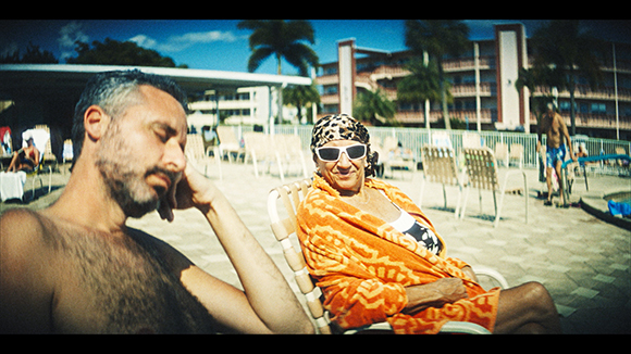 A man rests his head on his hand while a woman in sunglasses and an orange wrap sits in a lounge chair.