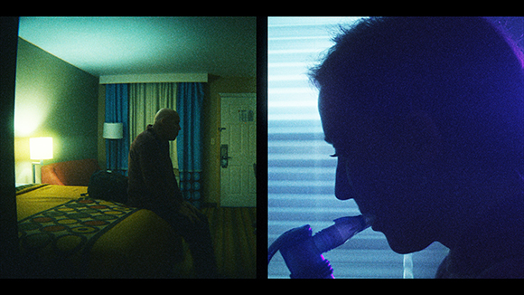 A man sits on a bed in a blue-lit room next to a silhouette of a person holding a microphone.