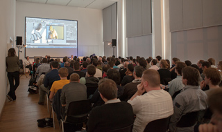 A large group of people seated in rows facing a large projection screen in a bright conference room.