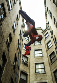 A superhero in a red and blue suit swings on a web between tall brick apartment buildings.