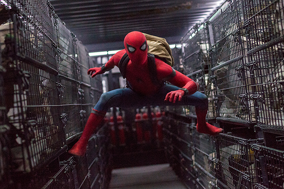 Spider-Man in a red and blue suit leaps through a dark corridor lined with metal storage cages.