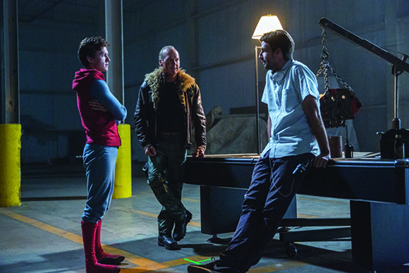 Three men stand together in a dark, industrial-looking room with a single overhead light.