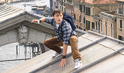 A young man in a plaid shirt crouches on a grey rooftop above a Venetian canal.