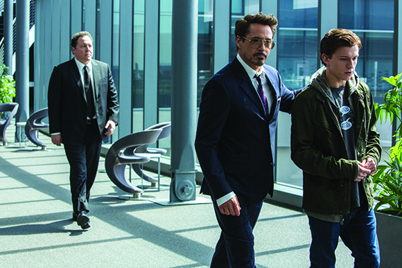 A man in a dark suit walks beside a younger man in a green jacket in a bright, modern glass hallway.