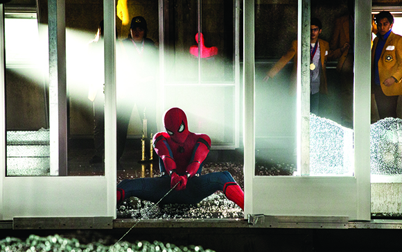 Spider-Man sits on a floor of broken glass in a smoky room as people watch through glass doors.