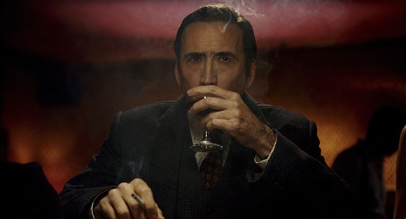 A man in a dark suit sits in a dimly lit room, drinking from a stemmed glass while holding a cigarette.