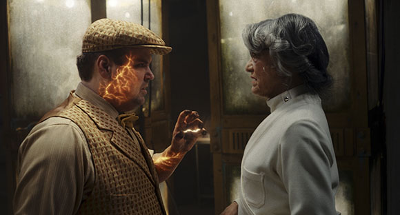 A man in a flat cap and vest stares at another man in a white high-collar shirt.