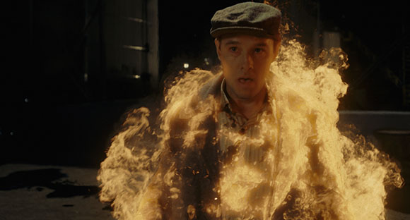 A man wearing a cap and shirt is engulfed in bright orange flames, with a look of shock on his face.