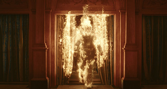 A figure completely covered in bright orange flames walks forward through a dark wooden doorway, with dark curtains on either