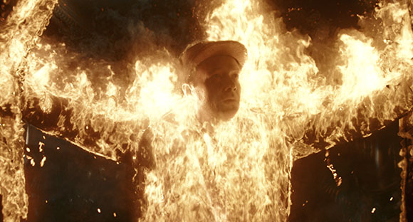 A man wearing a cap stands with arms outstretched while covered in bright orange flames.