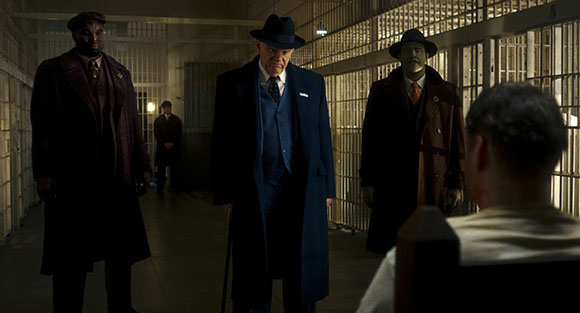 Three men in vintage coats and hats stand in a dimly lit prison hallway facing a person in the foreground.