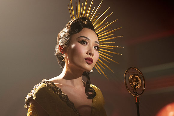 A woman in a gold dress and sunburst headpiece looks upward into a spotlight.