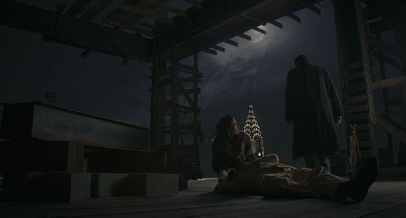 A man in a long coat stands over a woman lying on a wooden floor under a dark, cloudy night sky.
