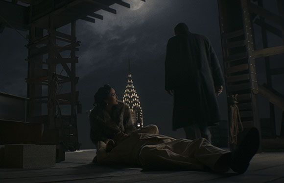 A woman in a fur coat kneels over a man lying on a rooftop at night, with the Chrysler Building visible in the background.