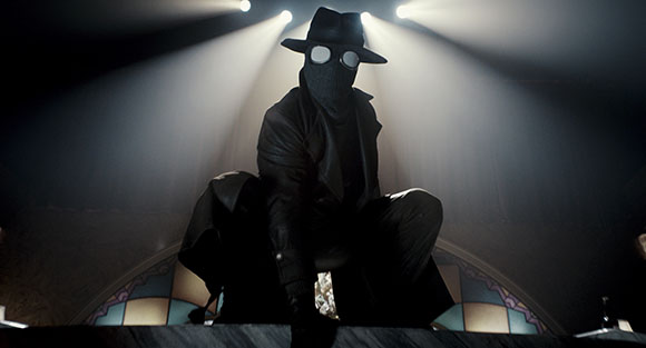 A person in a black mask, hat, and trench coat crouches under bright spotlights in a dark setting.