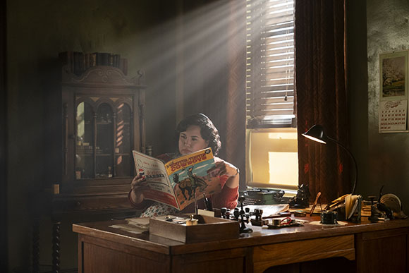 A woman sits at a wooden desk reading a magazine as sunlight streams through window blinds.