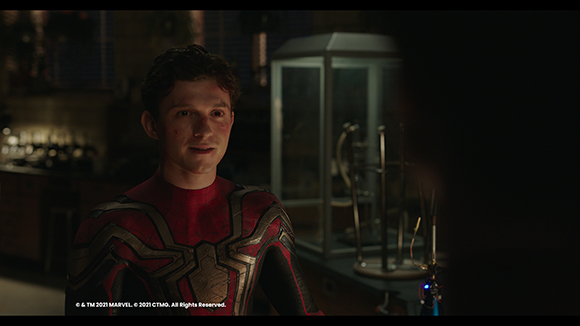A man with dark hair wearing a red and gold patterned superhero suit in a dimly lit room.
