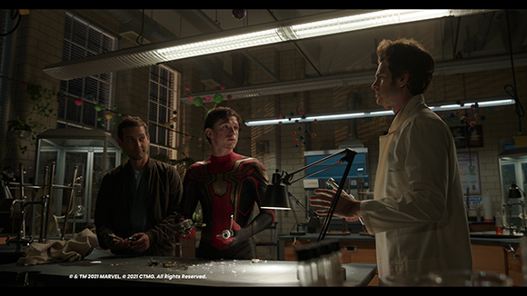 Three men stand in a dimly lit laboratory with scientific equipment and overhead fluorescent lights.