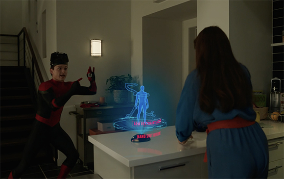 A man in a red and black suit gestures toward a blue holographic human figure on a white counter.