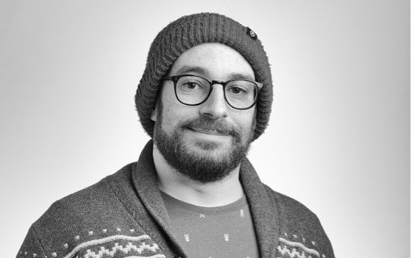 A man with a beard, glasses, and a knit beanie smiling slightly against a plain background.