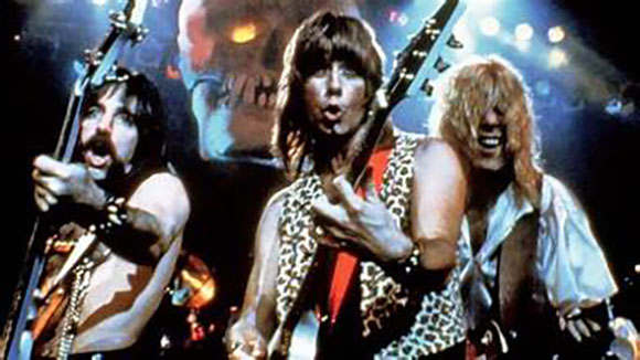 Three long-haired rock musicians performing on stage with a large skull graphic in the background.