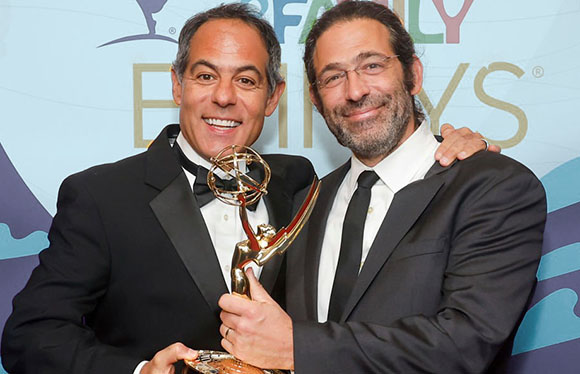 Two men in black suits smiling and holding a gold Emmy award.