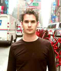 A man in a dark long-sleeved shirt stands on a busy city street with vehicles in the background.