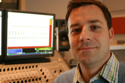 A man smiling in a studio with computer monitors and an audio mixing console in the background.